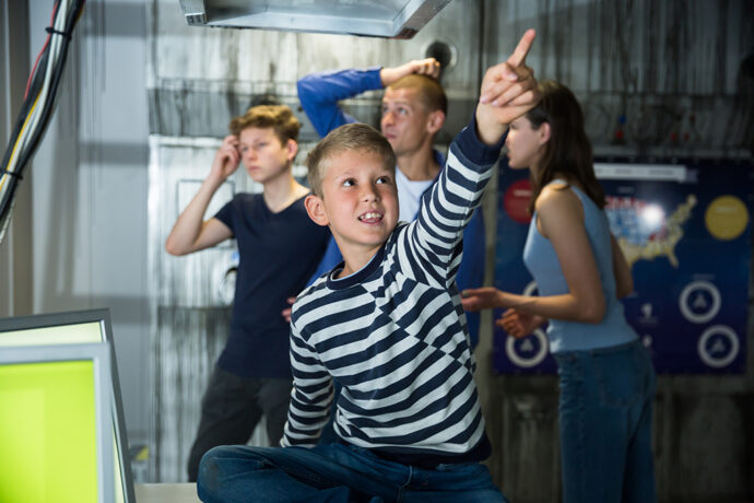 Emotional tween boy with his parents and older brother trying to get out of closed space of quest room stylized as abandoned bunker, enthusiastically pointing at something with his hand